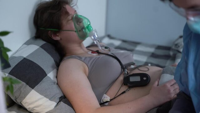 Covid 19, coronavirus. Doctor home visiting during quarantine, patient woman lying in bed with oxygen mask, medical worker doing electrocardiogram checking heart function. First aid, paramedicine.