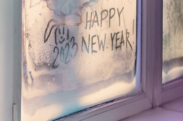 Drawing happy new year on a frozen window. The rabbit is the symbol of the Chinese New Year 2023. Happy New Year and Merry Christmas concept. Sunlight through frosty patterns. Selective focus
