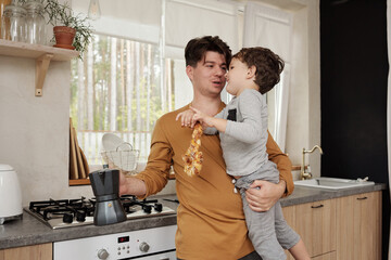 Mature man with coffee pot holding his little son chatting with him in kitchen horizontal medium shot