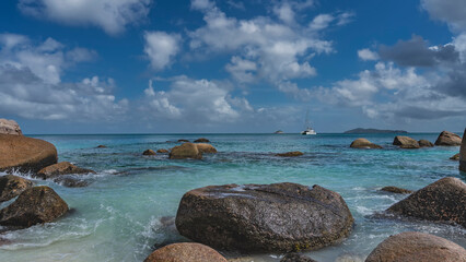 Picturesque granite boulders in the coastal waters of a tropical island. The waves of the turquoise ocean are beating against the rocks, foaming. The yacht is far away. Clouds in the blue sky. 