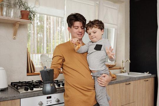 Caucasian man with coffee pot holding his little son interacting with him in kitchen horizontal medium shot