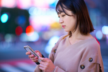 夜の繁華街でスマートフォンを見る若い女性
