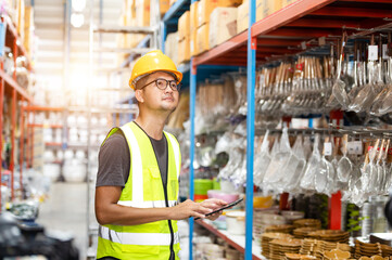Smart Asian engineer male worker wearing hard hat managing stock of household goods on shelves in...
