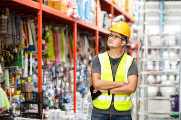 Smart Asian engineer male worker wearing hard hat managing stock of household goods on shelves in...