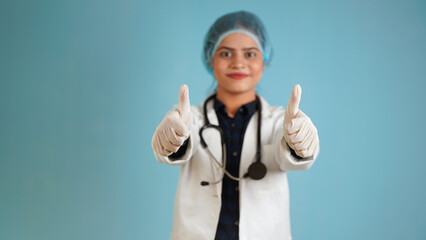 Portrait of a young female doctor gesturing thumbs up, Cheerful Asian Indian woman doctor in apron and stethoscope isolated over blue studio background