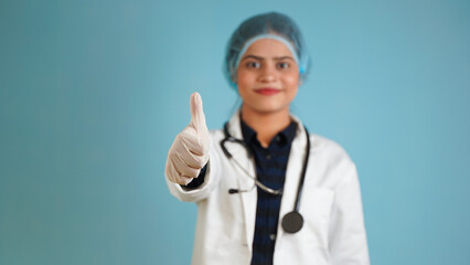 Portrait of a young female doctor gesturing thumbs up, Cheerful Asian Indian woman doctor in apron and stethoscope isolated over blue studio background