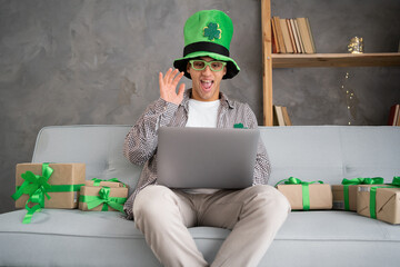 Young man in leprechaun hat video calling friends on laptop greeting family by webcam talk open St.Patrick's day gift box in virtual online chat meeting using computer sit at sofa on Patrick's day.
