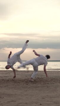 Two men in white clothes practicing capoeira on beach. Vertical video