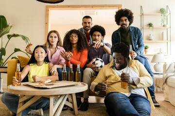 Excited group of friends watching diverse football game on TV, holding beer bottles and balls, eating pizza at home. Group of sports fans enjoying a television game
