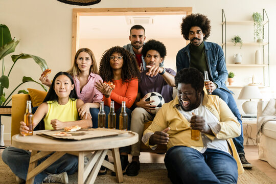 Excited Group Of Friends Watching Diverse Football Game On TV, Holding Beer Bottles And Balls, Eating Pizza At Home. Group Of Sports Fans Enjoying A Television Game