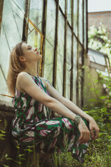 Leaning against window tiredly scenic photography. Picture of young woman with abandoned greenhouse on background. High quality wallpaper. Photo concept for ads, travel blog, magazine, article