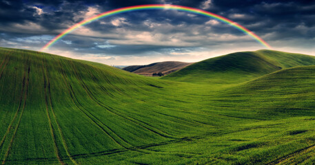 Obraz premium Scenic view of rainbow over green field. dramatic gray sky over a picturesque hilly field
