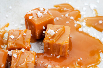 Salted caramel, a taste sensation. Coarse salt and caramel blocks covered in a sticky creamy salted caramel sauce on grey - obrazy, fototapety, plakaty