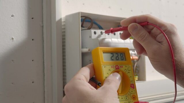 Work under voltage, electrician is checking serviceability of equipment, measuring voltage by yellow multimeter. Close up of a mans hands testing white switch box check voltage switchboard.