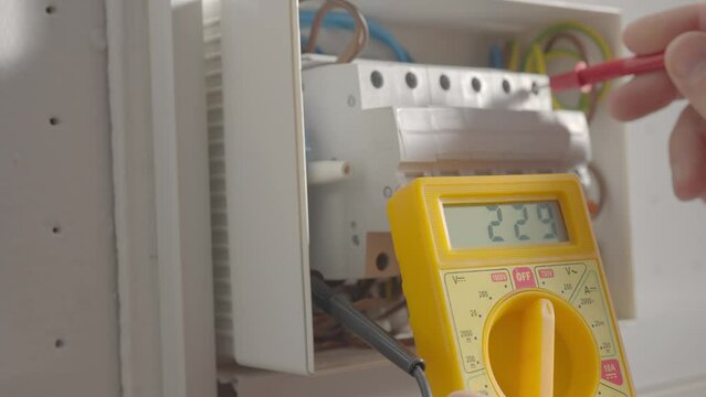 Work under voltage, electrician is checking serviceability of equipment, measuring voltage by yellow multimeter. Close up of a mans hands testing white switch box check voltage switchboard.