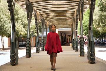 African American woman in a beautiful red party dress with large sleeves, walking and posing happily under an outdoor metal structure. Concept beauty, fashion, trend, empowerment, happiness.