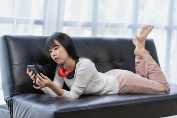 Obraz premium Relaxed happy asian woman lying on sofa at living room and using mobile phone at home lying down watching movie listening to music