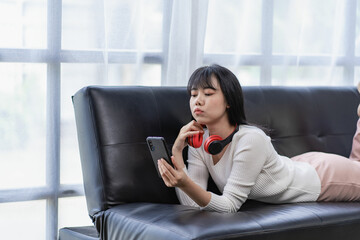 Relaxed happy asian woman lying on sofa at living room and using mobile phone at home lying down watching movie listening to music