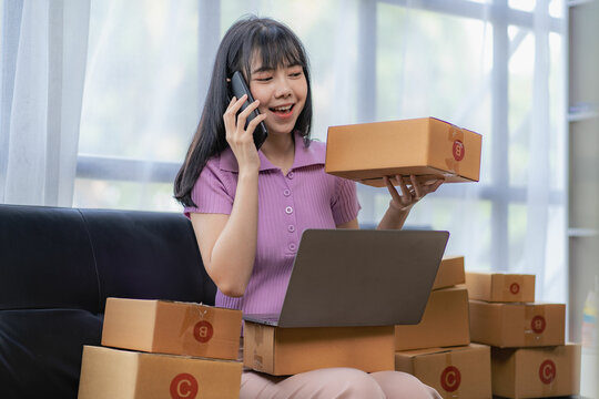 
Asian woman sitting on sofa and working at home, she is excited and smiling holding packaging box to write customer address in mailbox to send online sale items. The concept of SME business owners