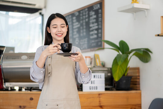 Beautiful Asian barista in gray apron holding cup of hot coffee customer service at bar counter