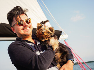 handsome person hugs his small dog yorkshire terrier on a sailing yacht during vacations, travel with pets concept