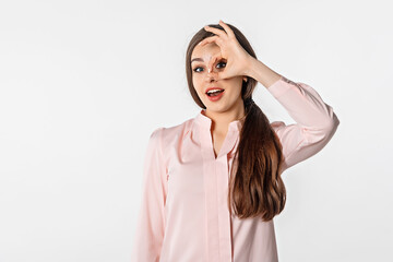 Excited young brunette woman is looking at camera, showing Ok sign and smiled, eye looking through fingers with happy face, standing against white studio background