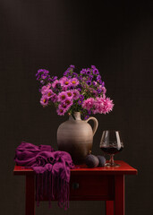 Bouquet of autumn flowers. Lots of lilac pink asters in a brown vase. Near a glass of red wine on a red wooden table. Two fresh figs. Dark pink shawl.Dark background. Still life.