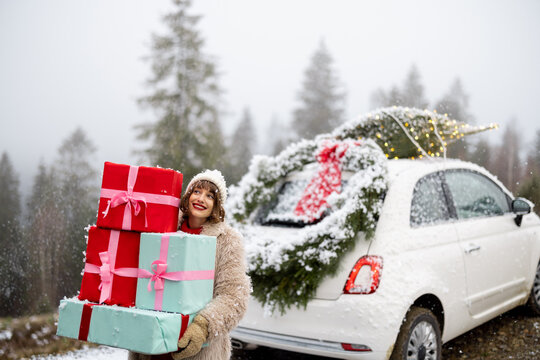 Woman Holds A Heap Of Gift Boxes While Standing By The Car Decorated With Christmas Wreath And Tree On A Rooftop, Traveling By Car In The Mountains On Snowy Weather. Concept Of Winter Holidays