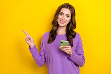 Photo of cheerful business influencer woman wear purple jumper hold phone finger point empty space advert isolated on yellow color background