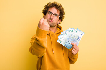 Young curly smart man holding euro banknotes isolated on yellow background showing fist to camera,...