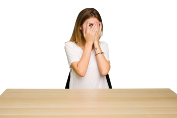 Young caucasian woman sitting on a chair in a desktop isolated blink through fingers frightened and nervous.