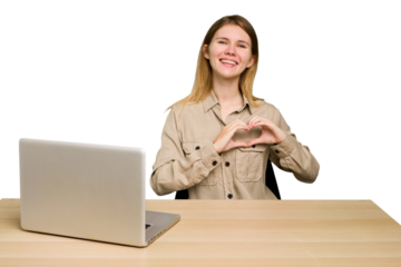 Young caucasian woman in a workplace working with a laptop isolated smiling and showing a heart shape with hands.