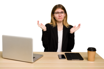 Young business caucasian woman working on her workplace cutout isolated doubting and shrugging shoulders in questioning gesture.