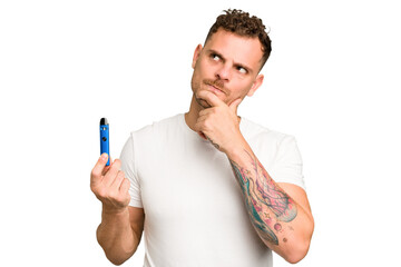 Young caucasian man holding a electronic cigarette isolated looking sideways with doubtful and...