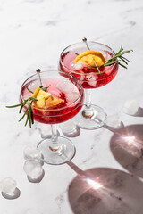 Two wineglass of Pomegranate Christmas iced cocktail with rosemary and lemon on white marble background isolated with clear shadows in harsh light. Xmas drink