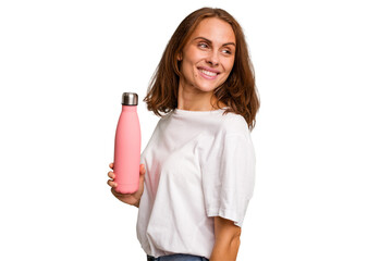 Young caucasian woman holding a pink thermo isolated looks aside smiling, cheerful and pleasant.