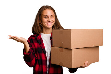 Young caucasian woman moving while picking up a box full of things isolated showing a copy space on a palm and holding another hand on waist.