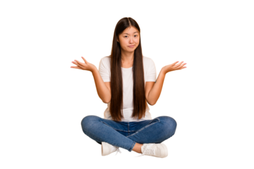 Young asian woman sitting on the floor cutout isolated doubting and shrugging shoulders in questioning gesture.
