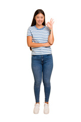 Young asian woman standing, full body cutout isolated winks an eye and holds an okay gesture with hand.