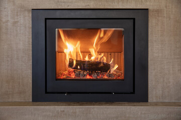 Burning fireplace, front view. Wood logs fireside, glass door, cast iron frame, marble wall background.