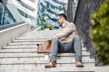 Obraz premium A businessman sits in front of the business center on the stairs and searching documents in his bag.