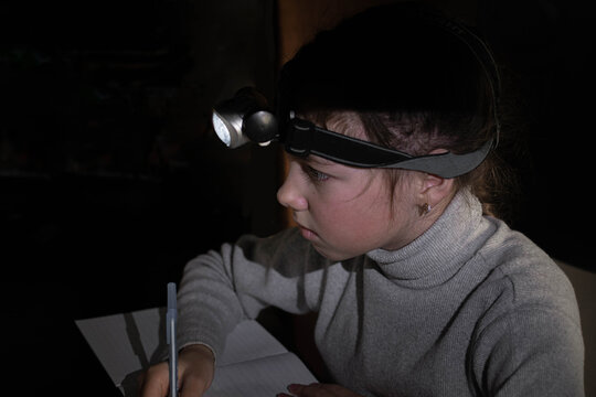 The Girl Learns Lessons In The Dark By Lighting Herself With A Headlamp. A Child Does Schoolwork When There Is No Electricity At Home.