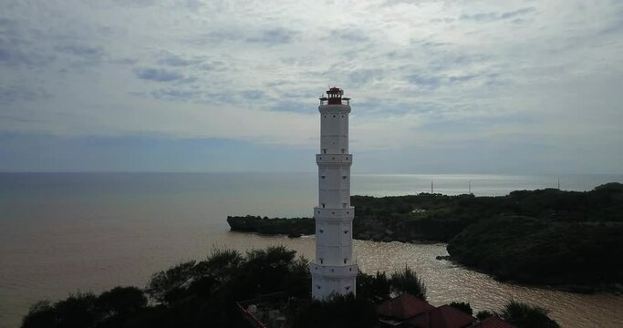 Tilt down drone shot of white lighthouse tower on the cliff borderer with sea in cloudy sky