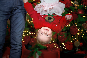 Happy smiling child on background of Christmas tree. little girl in New Year's outfit was turned upside down. Dad is holding child by legs.