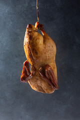 Smoked duck whole hanging upside down on hook on dark background.