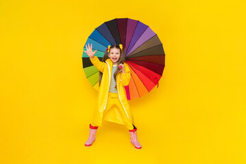 A little girl in a waterproof raincoat and rubber boots holds a beautiful multicolored umbrella in...