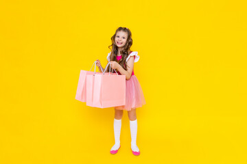 A little girl in a pink dress holds large shopping bags on a yellow isolated background. Spring shopping in the store, big promotions on goods.