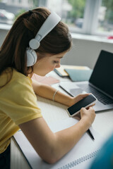 preteen girl in headphones holding smartphone with blank screen near blurred laptop