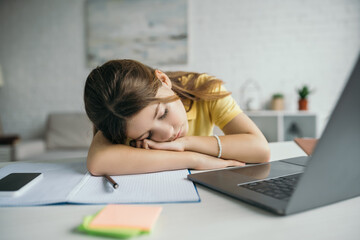 tired child sleeping near copybook and laptop with smartphone on table at home
