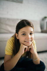 Fototapeta premium portrait of cheerful preteen girl sitting with hands near face and looking at camera